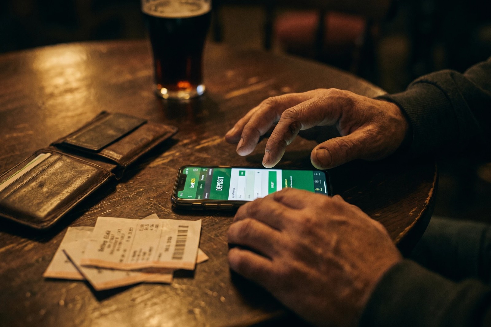 A man at a dark pub table tapping the deposit screen of a sports betting app on his phone, beside an open wallet, betting slips and a pint of beer